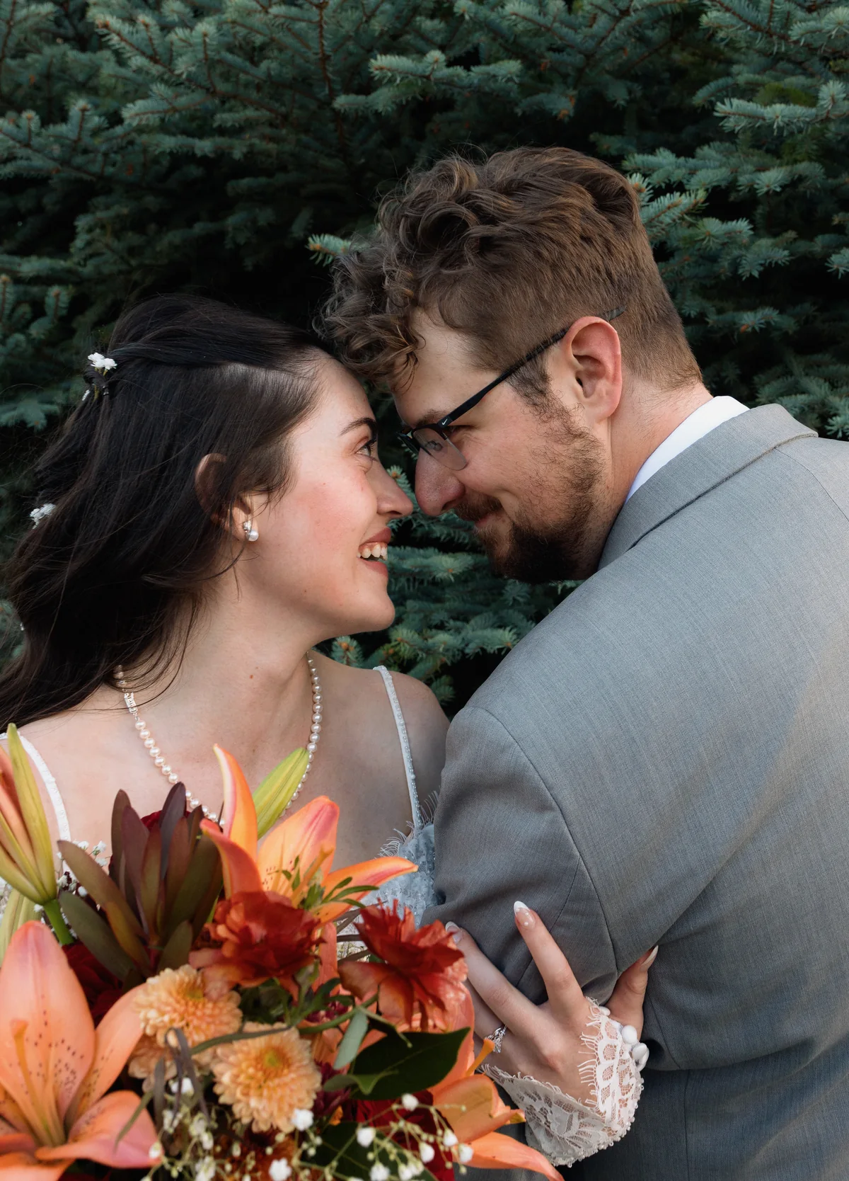 Bride and groom forehead to forehead and smiling in front of blue spruce, her orange lily bouquet in the foreground