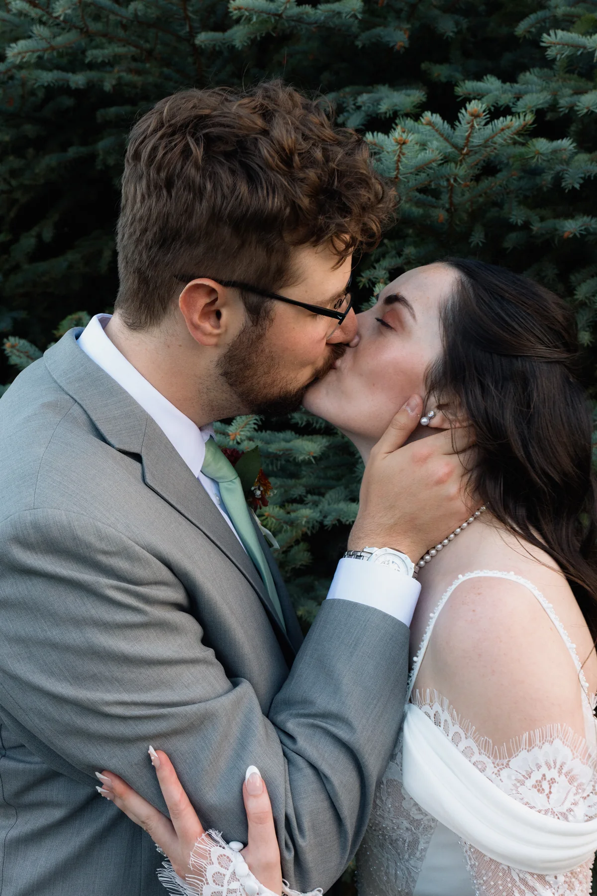 Groom kissing the bride, his hand on her cheek, in front of evergreens